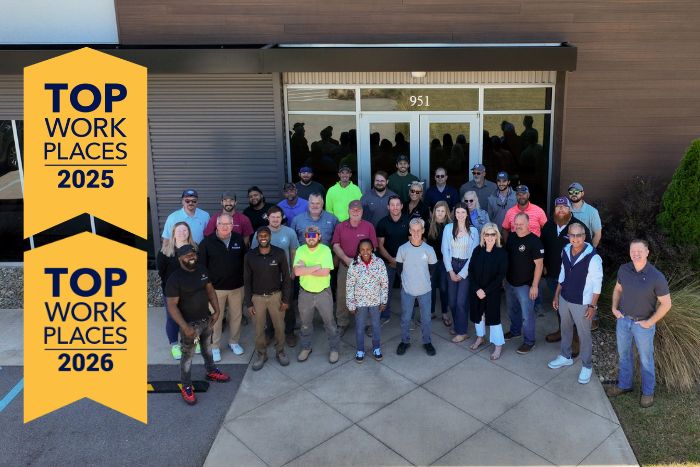 Group photo of diverse employees standing outside a building, beside a tall yellow banner reading Top Work Places 2025 and 2026.