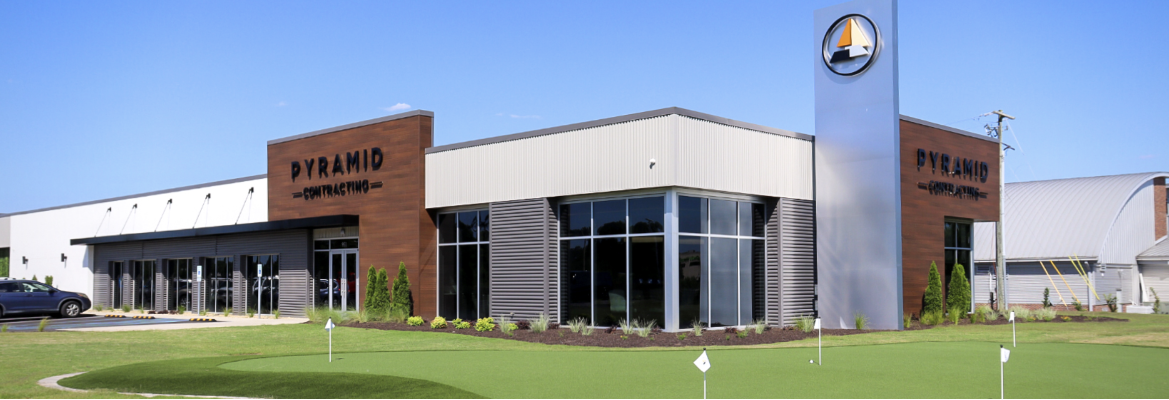 Pyramid Contracting Headquarters - Pyramid Contracting Exterior view of Pyramid Contracting office with wood-paneled façade, glass storefront, a tall sign tower bearing the logo, and a landscaped lawn with a putting green in front.