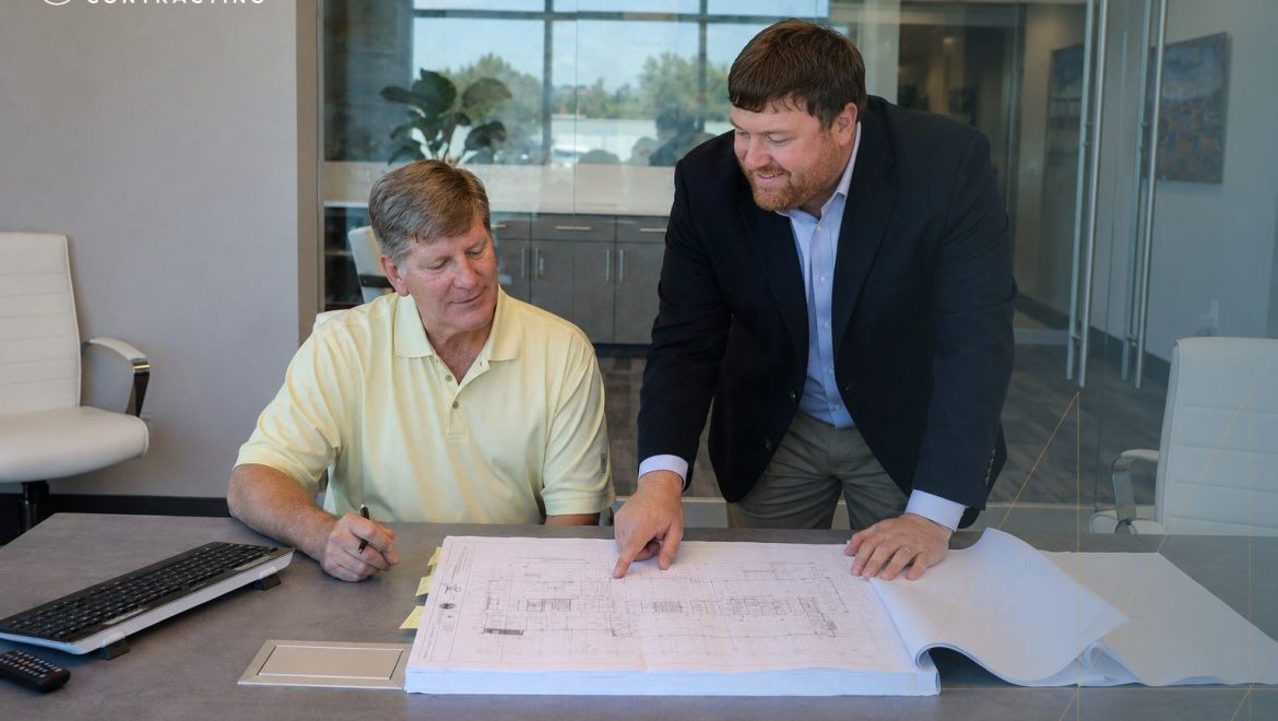Two men in an office review architectural blueprints spread on a table, with a laptop nearby.