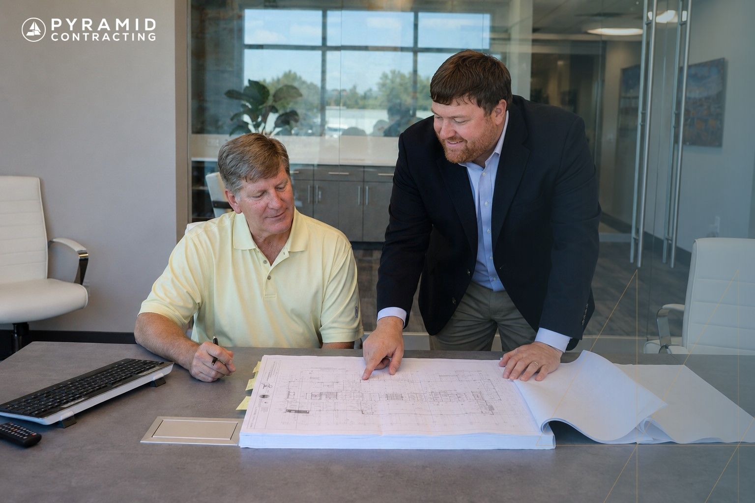Two men in an office review architectural blueprints spread on a table, with a laptop nearby.