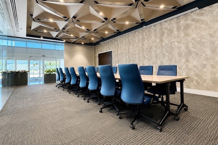 Long conference table with blue ergonomic chairs in a modern meeting room with geometric ceiling panels and natural light.